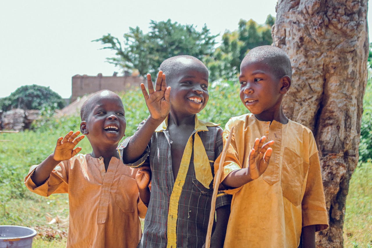 services-02 Three happy children waving outside in a vibrant Nigerian village setting.